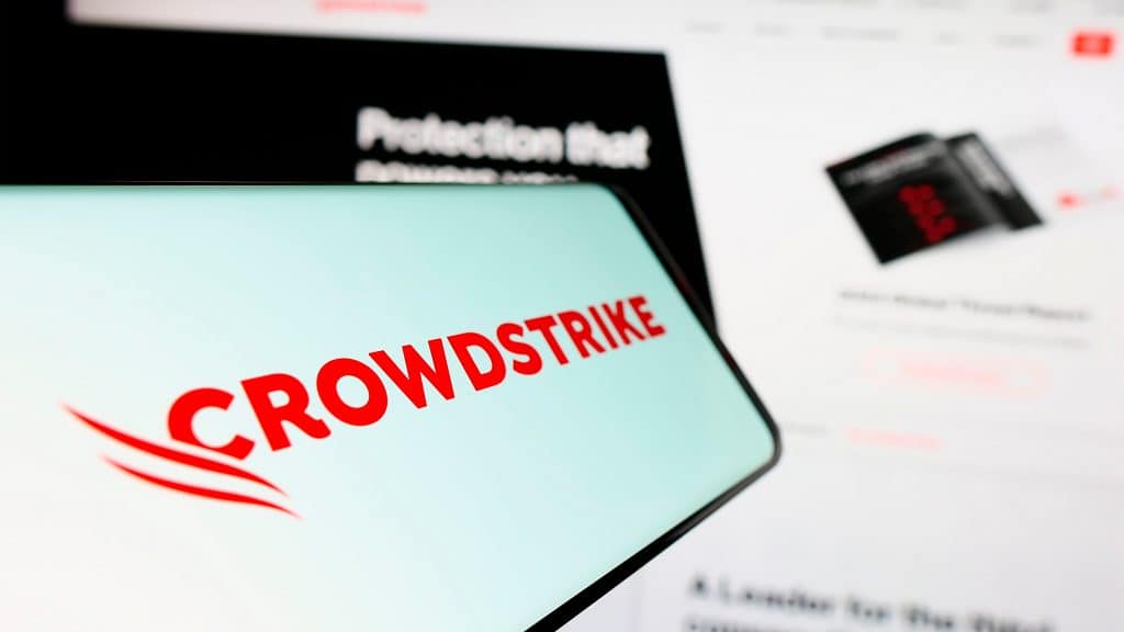 Celular com logotipo da Crowdstrike na tela e, ao fundo, computador com gráfico da empresa aberto