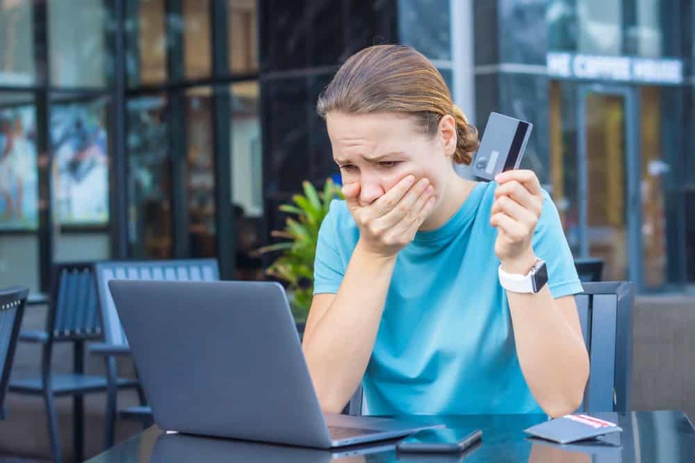 Mulher com uma mão na boca, com expressão de desespero, enquanto segura um cartão de crédito com a outra mão e olha para a tela de um notebook