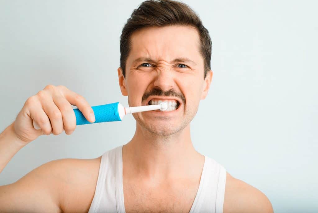 Retrato de um homem bonito escovando os dentes de forma persistente e enérgica com uma escova de dentes elétrica moderna, olhando para a câmera dentro de casa. A rotina diária de higiene bucal e clareamento dental de um jovem.