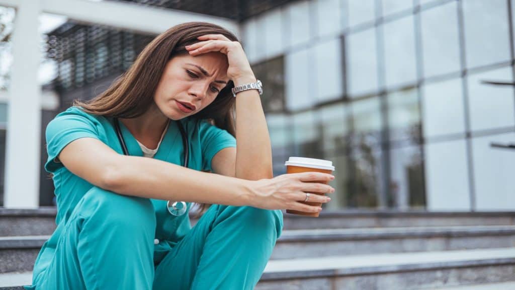 
mulher médica moderna estressada de uniforme com estetoscópio e xícara de café do lado de fora perto da clínica. Enfermeira se sentindo mal com uma dor de cabeça forte enquanto trabalha dentro do hospital.