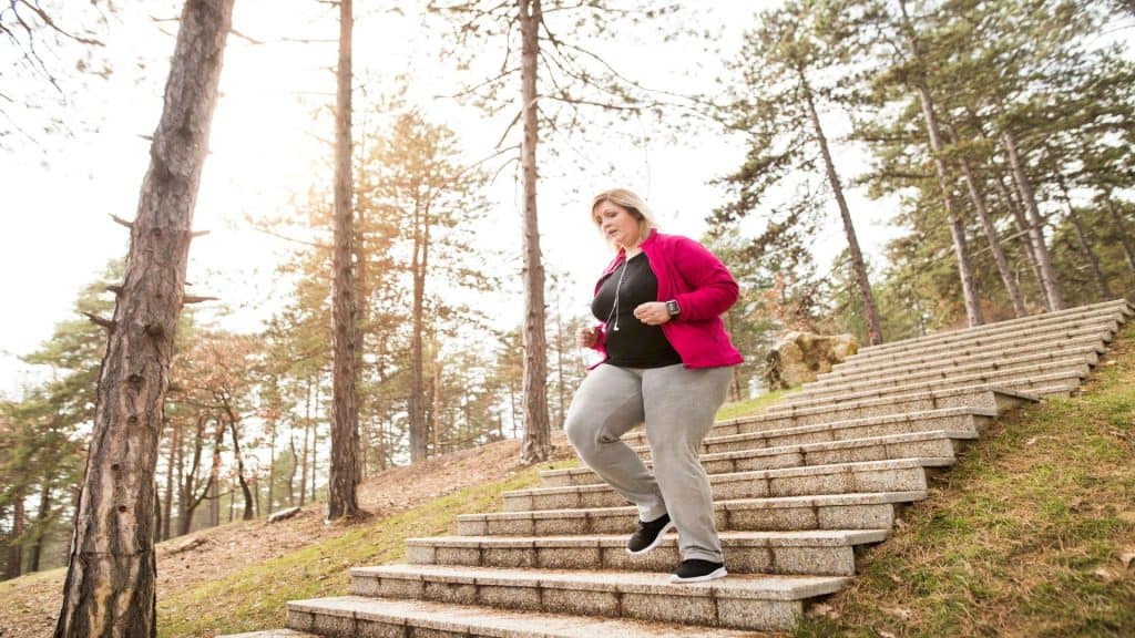 Mulher acima do peso correndo em parque da cidade, descendo as escadas. Exercitando-se ao ar livre para pessoas com obesidade.