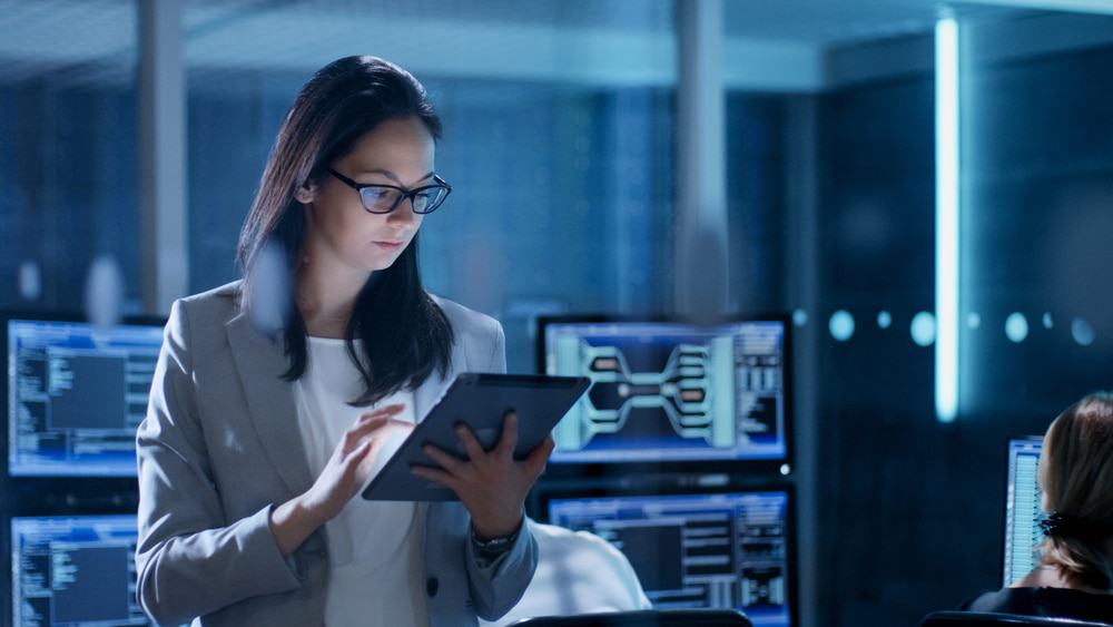 Imagem mostra uma mulher segurando um tablet dentro de uma sala com diversos computadores